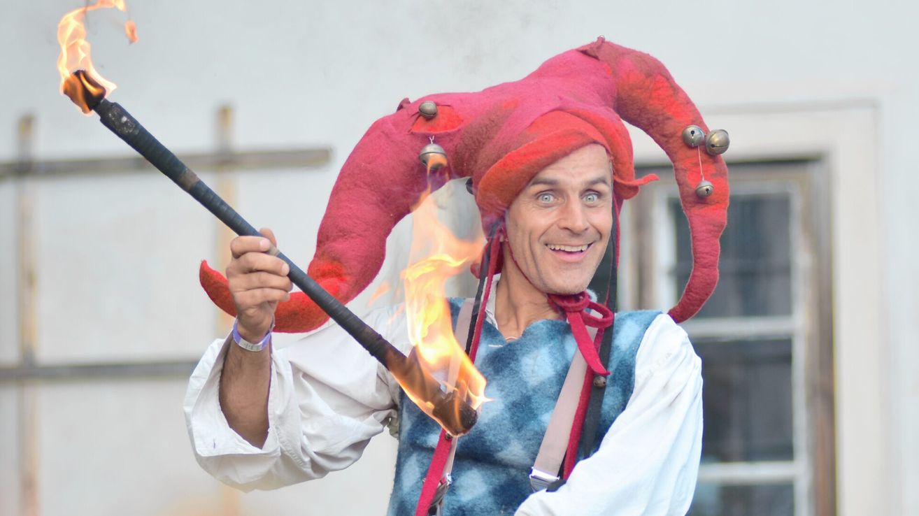 Ein Mann in einem Narrenkostüm, mit einem roten Hut mit Glocken und einer brennenden Fackel in der Hand, lächelt für die Kamera.