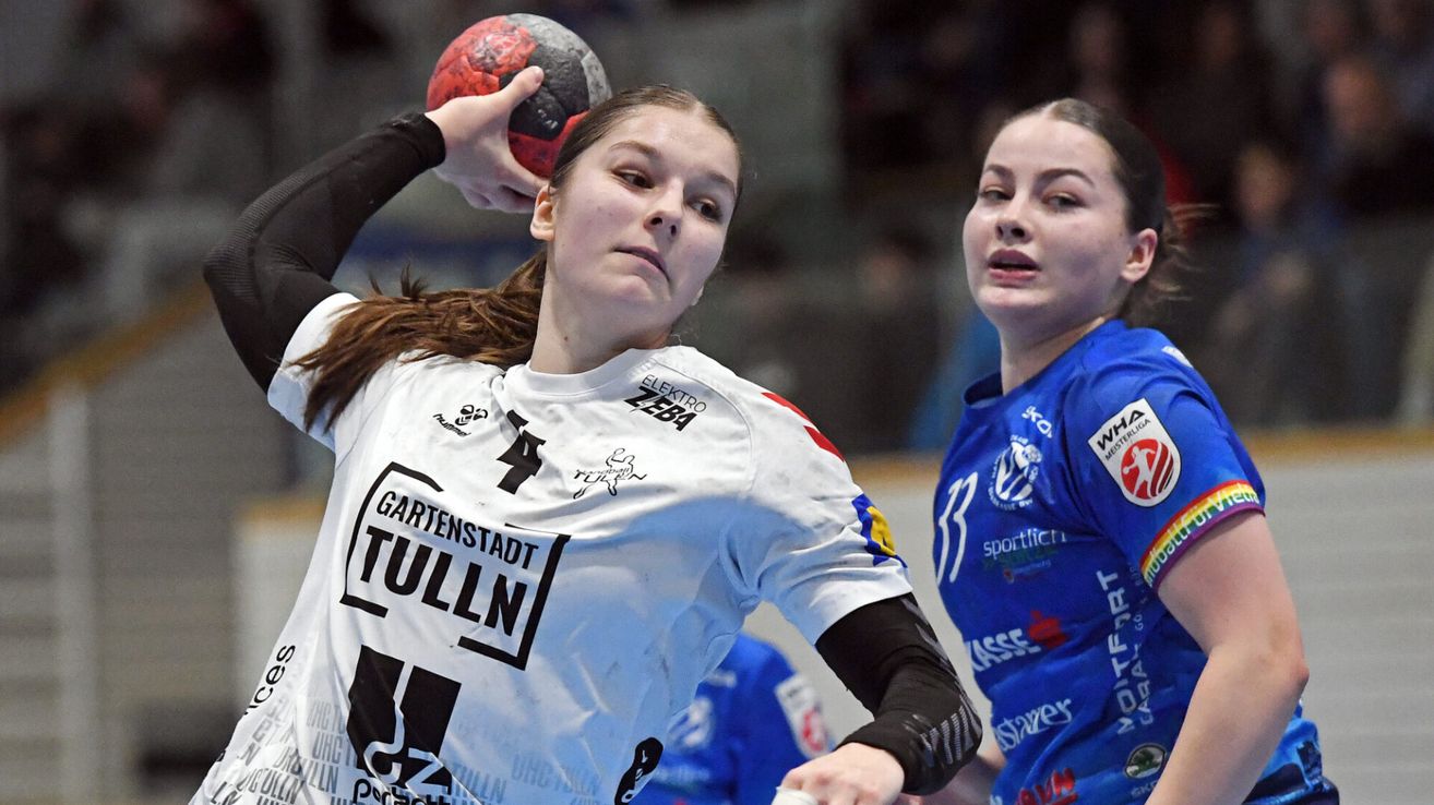 Zwei Frauen spielen Hallenhandball. Eine Spielerin, die ein weißes Trikot mit dem Wort Tulln trägt, hält einen Ball. Die andere Spielerin trägt ein blaues Trikot.