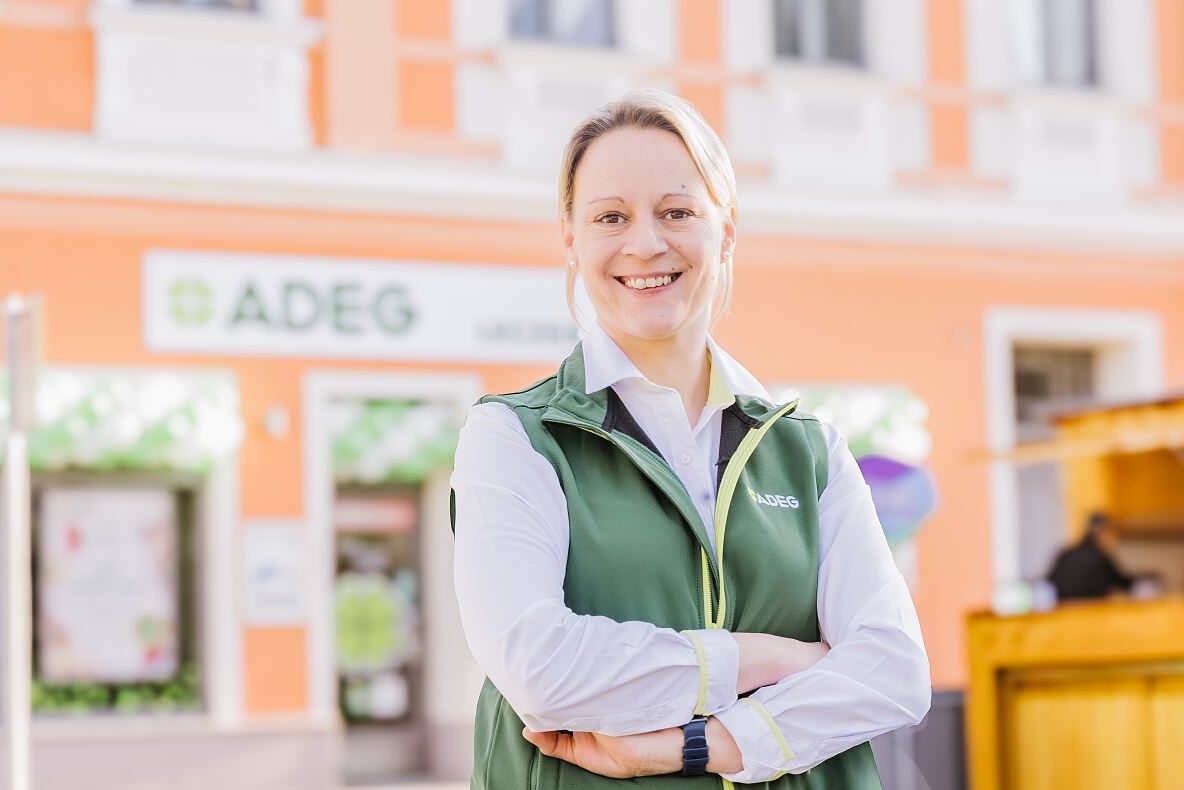 Eine Frau in einer grünen Weste steht mit verschränkten Armen vor einem orangefarbenen Gebäude mit dem Wort ADEG darauf.