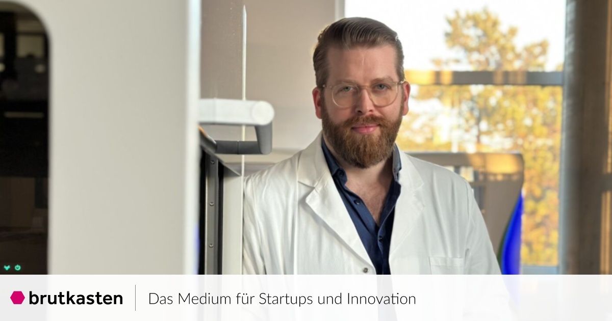 Ein Mann in einem Laborkittel und Brille steht in der Nähe einer Tür, möglicherweise in einem Laborumfeld. Der Hintergrund ist unscharf mit einem Fenster und Bäumen. Das Bild hat einen Wasserzeichen mit der Aufschrift 'Das Medium für Startups und Innovation.'