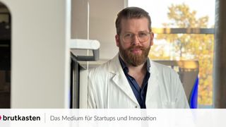 Ein Mann in einem Laborkittel und Brille steht in der Nähe einer Tür, möglicherweise in einem Laborumfeld. Der Hintergrund ist unscharf mit einem Fenster und Bäumen. Das Bild hat einen Wasserzeichen mit der Aufschrift 'Das Medium für Startups und Innovation.'