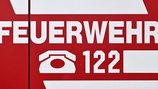 Rotes Schild mit weißem Text, der 'FEURWEH 122' und ein weißes Telefonsymbol neben der Nummer 122 zeigt.