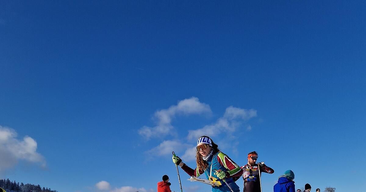 Eine Gruppe von Skifahrern in bunter Ausrüstung steht auf einem verschneiten Berg. Eine Frau hält eine Karte, während ein Mann seine Brille justiert. Sie sind von einem klaren blauen Himmel umgeben.