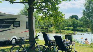 Ein Wohnmobil steht am See. Ein Fahrrad, zwei Stühle und ein Tisch sind auf dem Gras aufgestellt.