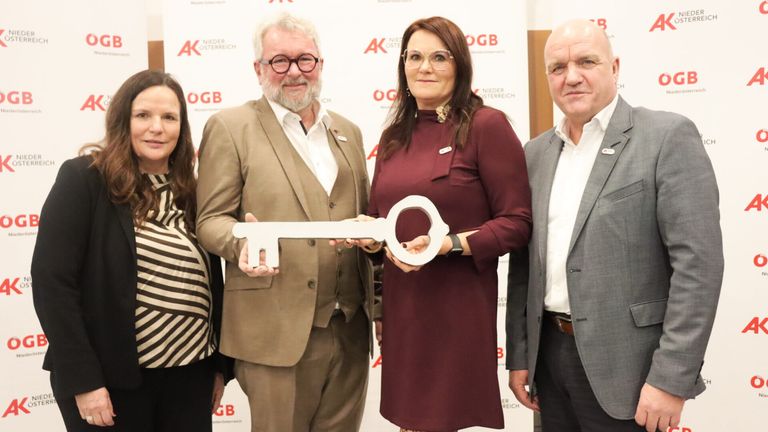 Vier Personen stehen gemeinsam lächelnd für ein Foto, wobei eine Frau einen Schlüssel hält. Der Mann links trägt eine Brille und einen Anzug, während die Frau rechts ein Weinrotkleid trägt.