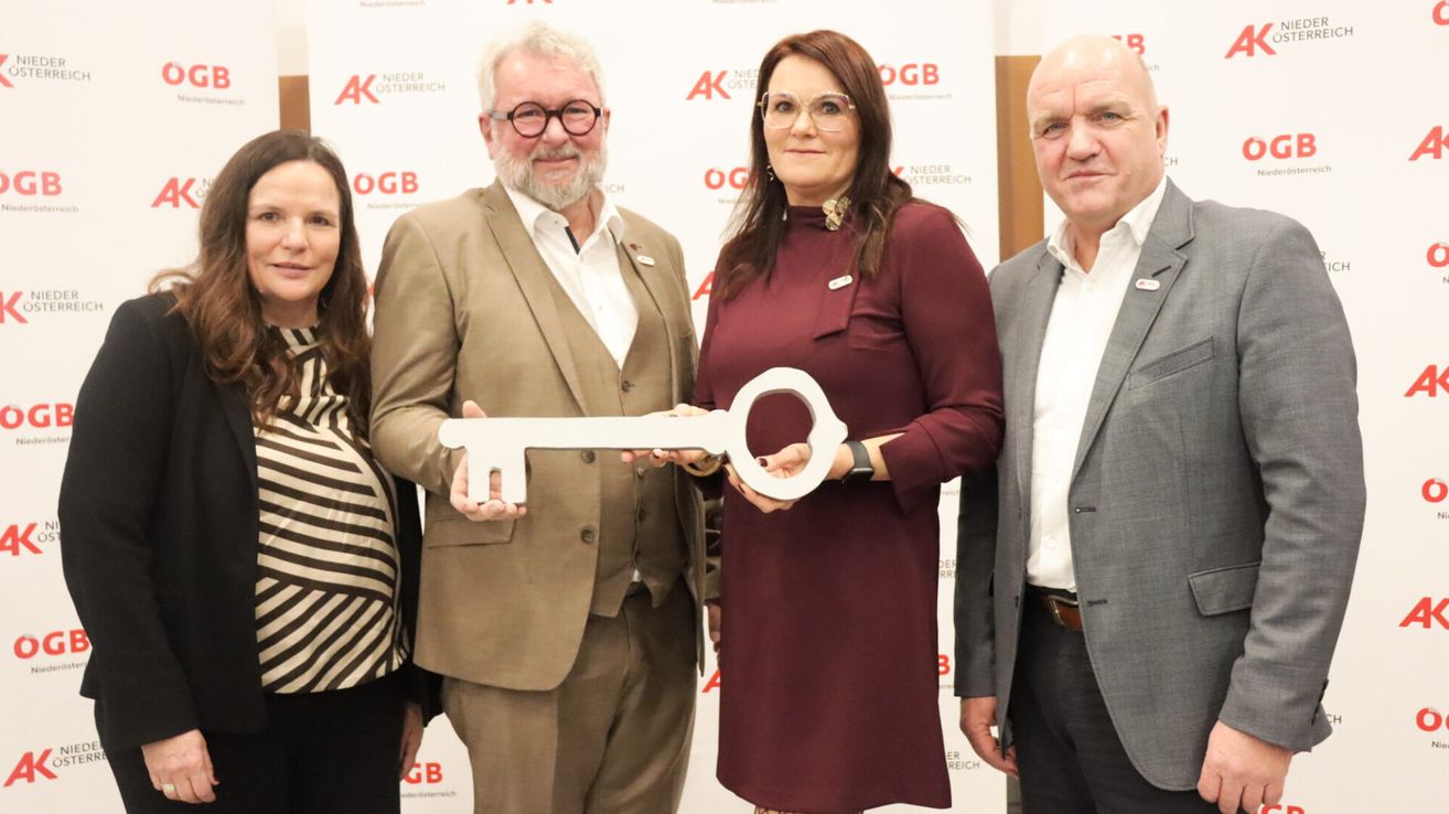 Four individuals stand together, smiling for a photograph, with a woman holding a key. The man on the left wears glasses and a suit, while the woman on the right wears a maroon dress.