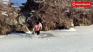 Ein Mann steht in eisigem Wasser in der Nähe eines schneebedeckten Ufers, trägt einen Hut und schwarze Shorts, mit trockenen Zweigen dahinter.