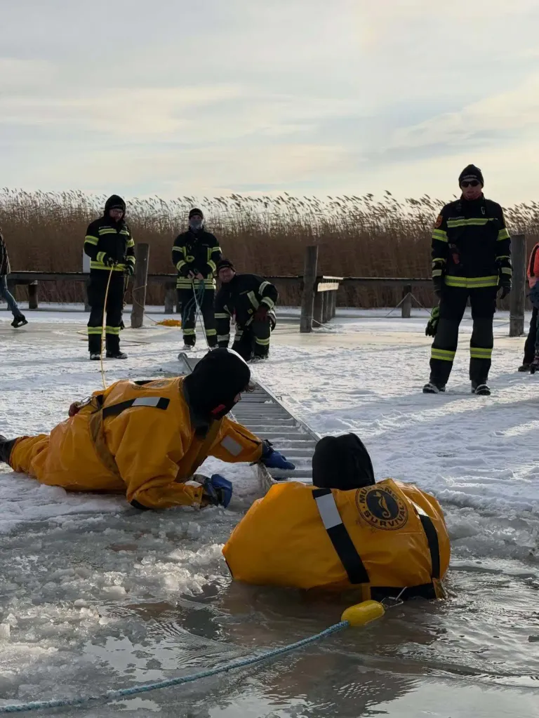 Feuerwehrleute führen ein Wasserrettungstraining in einer verschneiten Umgebung durch. Eine Person liegt im Wasser, während andere mit Seilen bereitstehen.