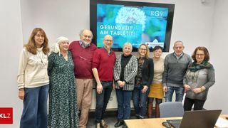 Eine Gruppe von Menschen steht vor einer Beamerleinwand, auf der Gesundheit am Tablet steht. Sie lächeln und scheinen für ein Foto zu posieren.