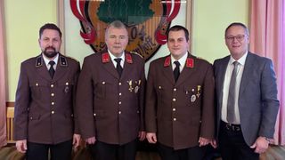 Three men in military uniforms stand next to each other, smiling, in front of a wall with a logo.
