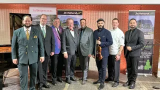 Neun Männer in Anzügen posieren für ein Foto mit einem Violinisten, vor Bannern für 'tulbingerkogel' und 'Dr. Richard JUF' stehend.