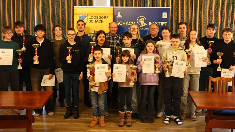 Eine Gruppe von Menschen steht zusammen und hält Pokale. Sie scheinen eine Schachveranstaltung zu feiern. Hinter ihnen zeigen Banner die Wörter 'Loosdof Schach' und 'IM Stift'.