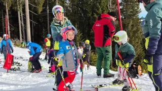 Eine Gruppe von Menschen skifahrer auf einem verschneiten Hang, umgeben von Tannen. Einige tragen Helme und halten Skistöcke. Eine Frau unterrichtet ein Kind.