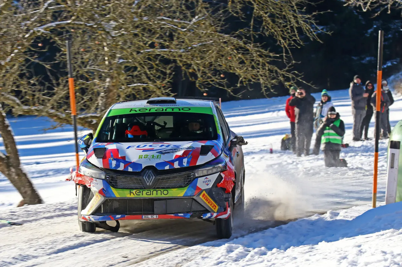 A rally car with the name Keramo on the side is driving on a snowy road. The car is covered in snow. A few people are standing behind the car.