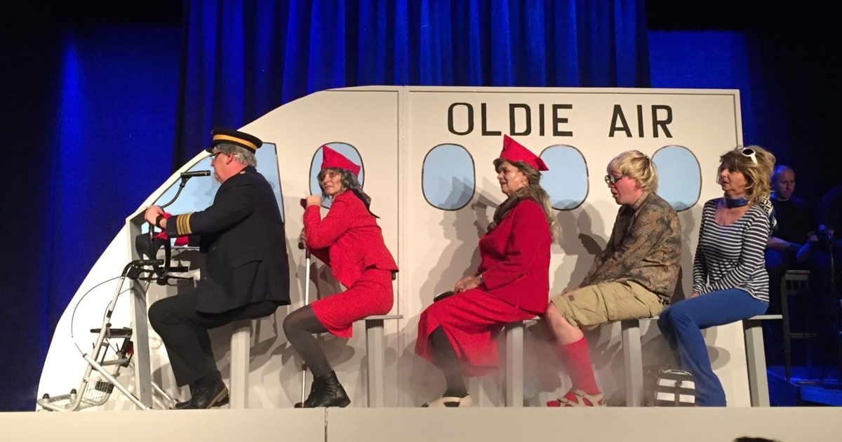 Vier Personen in roten Kostümen und Hüten sitzen auf Hockern vor einem nachgebauten Flugzeug, das Oldie Air beschriftet ist.