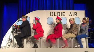 Vier Personen in roten Kostümen und Hüten sitzen auf Hockern vor einem nachgebauten Flugzeug, das Oldie Air beschriftet ist.