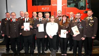Eine Gruppe von Feuerwehrleuten in Uniform, die Urkunden halten, posiert vor einem roten Feuerwehrauto. Dazu gehören zwei Frauen in der Mitte und mehrere Männer an den Seiten.