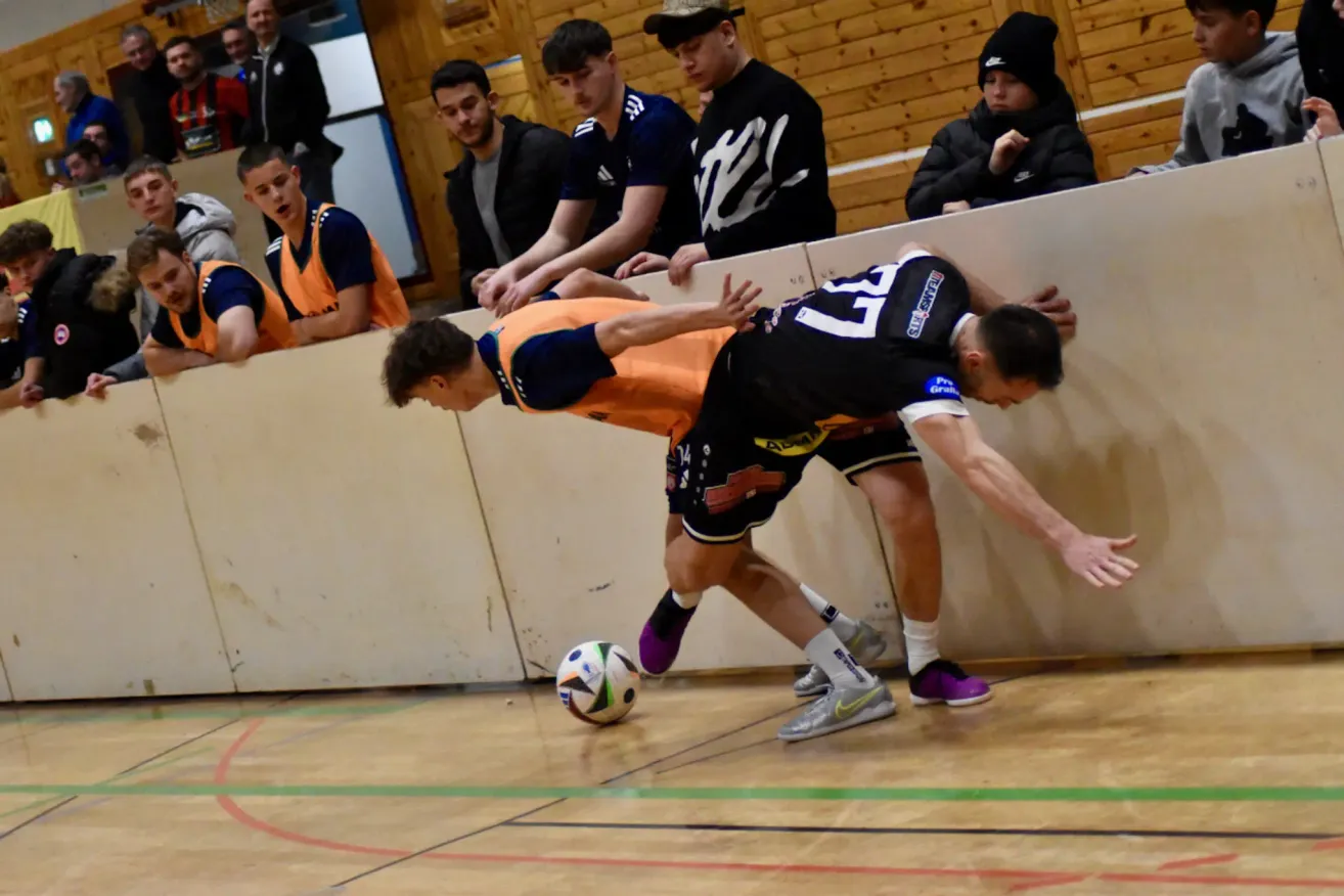 Zwei Spieler spielen Fußball in einer Halle. Einer trägt ein orangefarbenes Trikot, der andere ein schwarzes. Sie beugen sich über den Ball. Dahinter beobachten mehrere Personen.