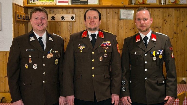 Drei Männer in Militäruniformen stehen vor einer Holzwand, einer lächelt und die anderen beiden haben ernste Gesichtsausdrücke.