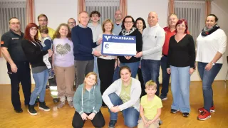 Eine Gruppe von Menschen steht in einem Raum und hält einen Scheck von der Volksbank über 500 Euro. Sie lächeln für die Kamera, einige knien und andere stehen. Der Raum hat Holzböden und Vorhänge.