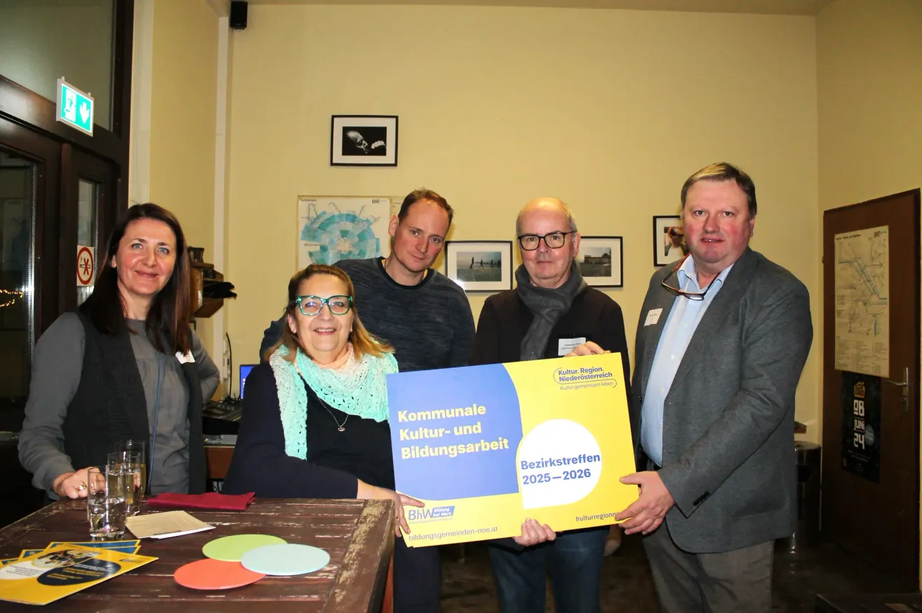 Five people stand in a room with a yellow poster. A woman holds the poster while smiling. Behind her, a man and a woman are looking at the poster. Another two men are standing next to them. The wall has picture frames and a map.