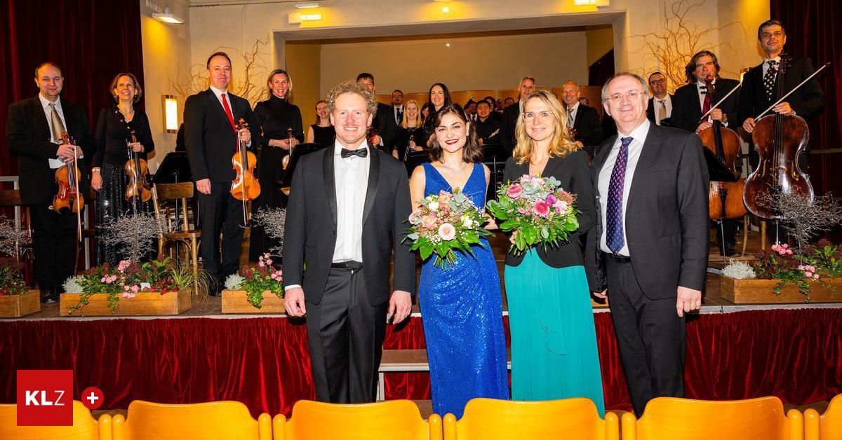 Eine Gruppe formell gekleideter Personen, darunter Musiker, steht auf der Bühne, ein Violinist hält sein Instrument und zwei Frauen halten Blumensträuße.