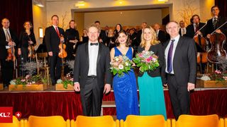 Eine Gruppe formell gekleideter Personen, darunter Musiker, steht auf der Bühne, ein Violinist hält sein Instrument und zwei Frauen halten Blumensträuße.