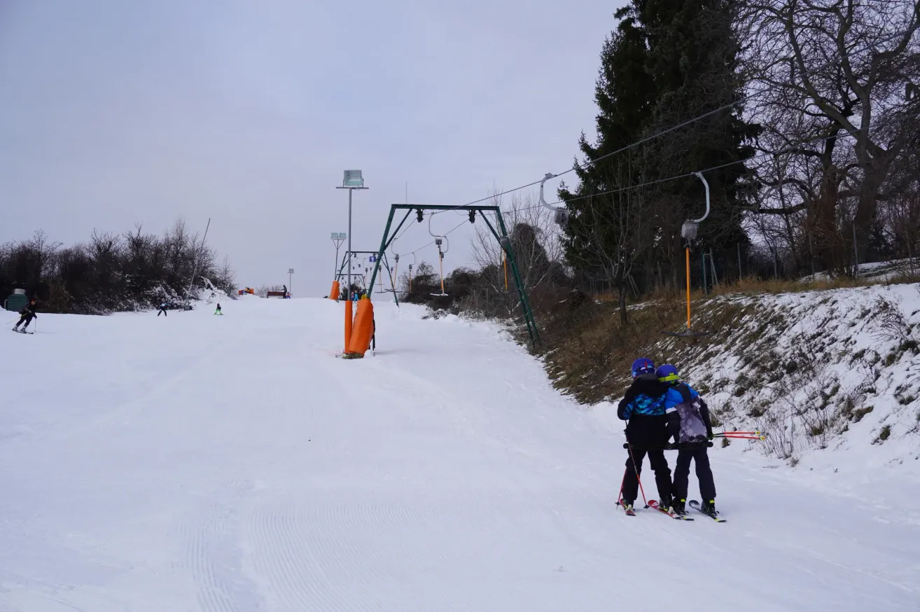 Two skiers, one holding a child, stand on a snowy slope with ski lifts above. Trees and plants are around. Other skiers are in the distance.