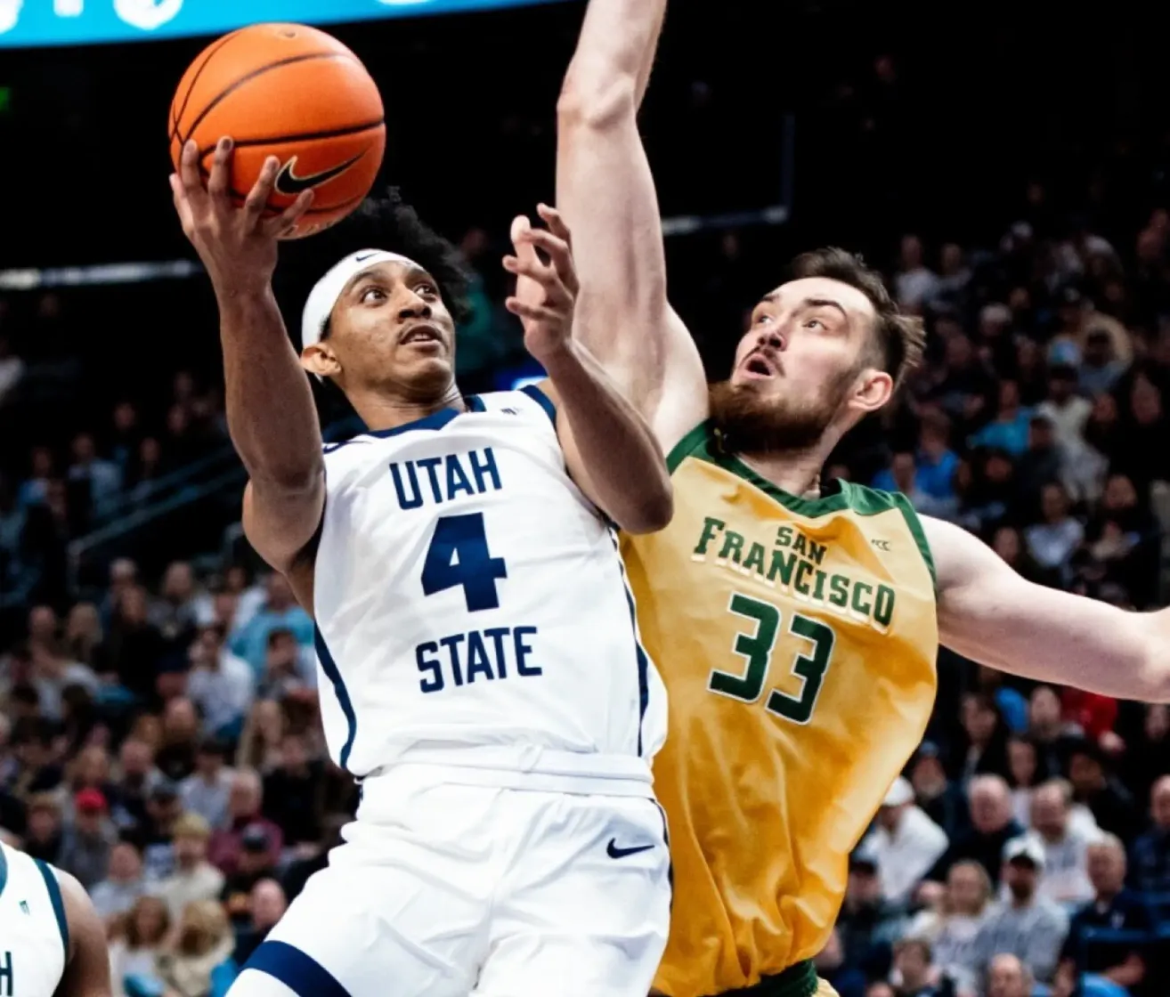 Während eines Basketballspiels versucht ein Spieler von Utah State mit der Trikotnummer 4 zu werfen, während er von einem Spieler aus San Francisco in einem gelben Trikot mit der Nummer 33 blockiert wird.