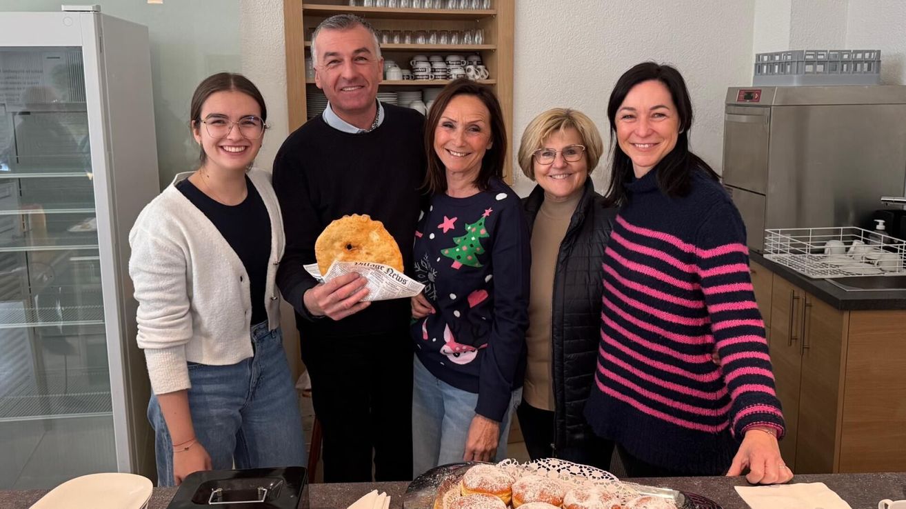 Five individuals stand together in a room, smiling for a photo. A man holds a pie wrapped in newspaper. Two women wear Christmas sweaters, one with a tree design. A woman in glasses and another in a striped sweater stand on the right. Behind them, a shelf holds plates and cups.