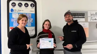 Drei Personen stehen vor einer Wand. Eine Frau links und eine Frau in der Mitte mit einem Zertifikat. Der Mann rechts zeigt auf das Zertifikat.