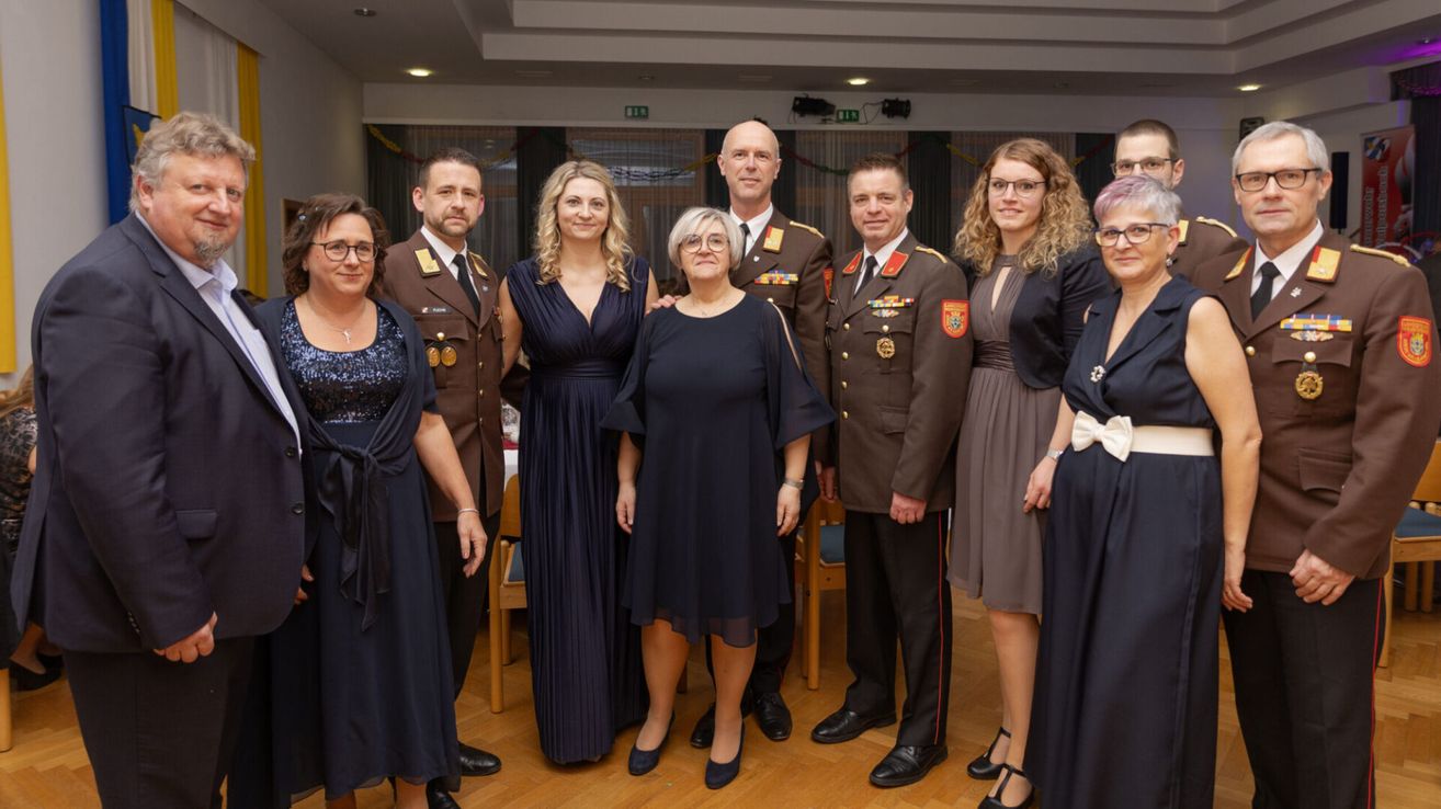A group of formally dressed individuals are posing for a photo, including military officers and civilians, standing in a well-lit room with wooden floors and decorative lights.