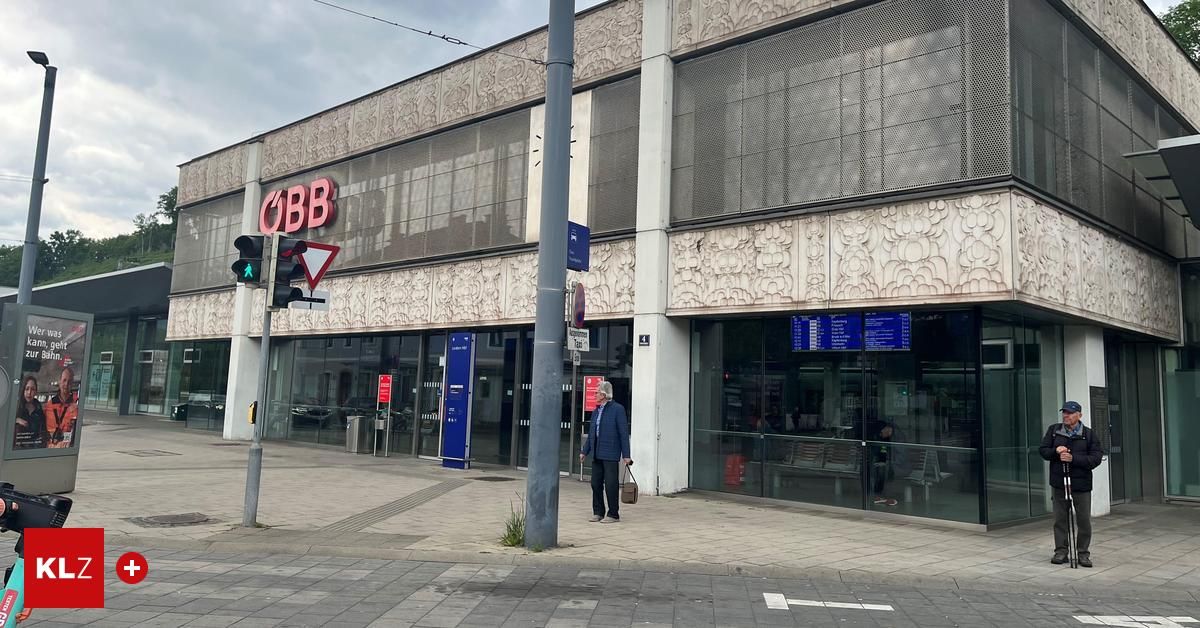 Ein älterer Mann in einem Mantel und mit einer Handtasche geht vor einem Gebäude eines Verkehrsknotenpunkts. Das Gebäude hat Glasfenster und ein Schild, das 'QBB' steht.