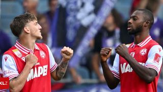 Zwei männliche Fußballspieler feiern ein Tor, tragen rote Trikots mit weißen Streifen und Tattoos und stehen vor einer jubelnden Menge.