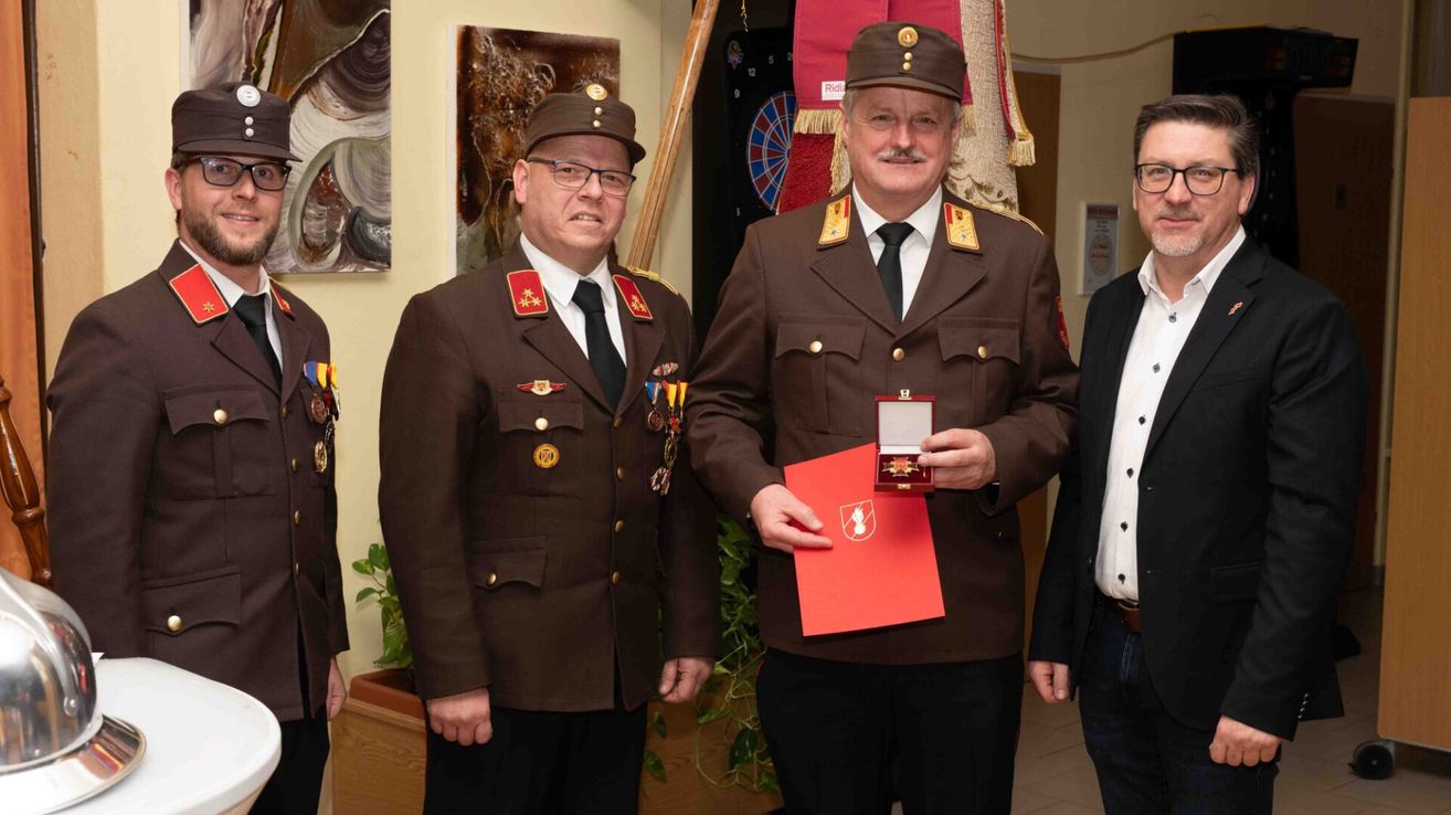 Zwei Männer in Militäruniformen stehen nebeneinander. Der Mann auf der rechten Seite hält eine rote Mappe und eine Medaillendose. Hinter ihnen steht ein Mann in einem Anzug und eine Wand mit Gemälden und einem Dartboard.
