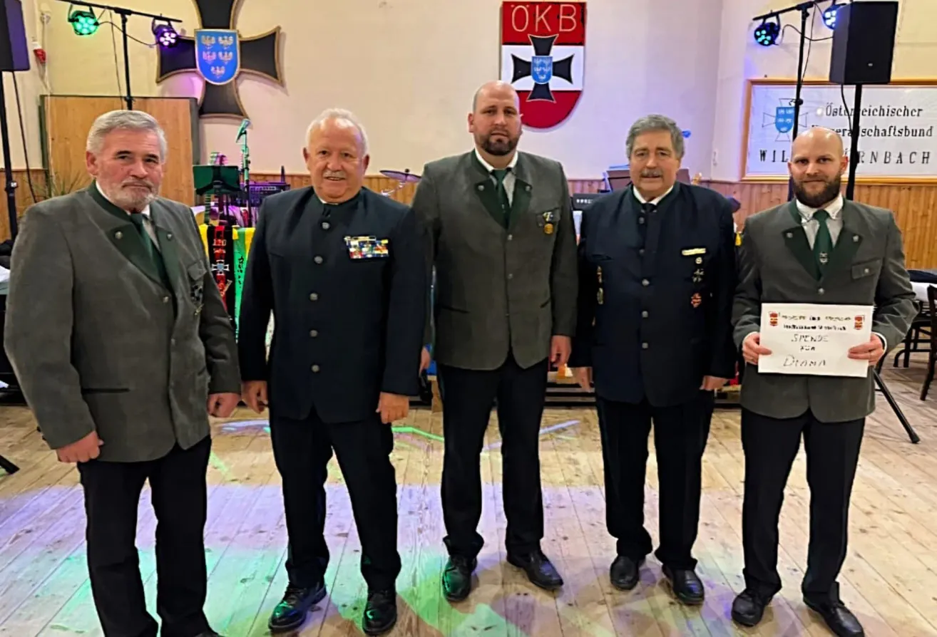 Fünf Männer in formeller Kleidung stehen in einem Raum mit Holzboden. Sie lächeln und schauen in die Kamera. Hinter ihnen befindet sich eine Wand mit einem gerahmten Bild und einem Schild.
