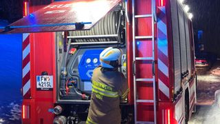 Ein Feuerwehrmann in voller Ausrüstung, mit Helm, arbeitet an den Geräten in einem roten Feuerwehrwagen mit einer Leiter an der Seite. Der Wagen hat ein Kennzeichen mit der Aufschrift 'FW 421SL.'