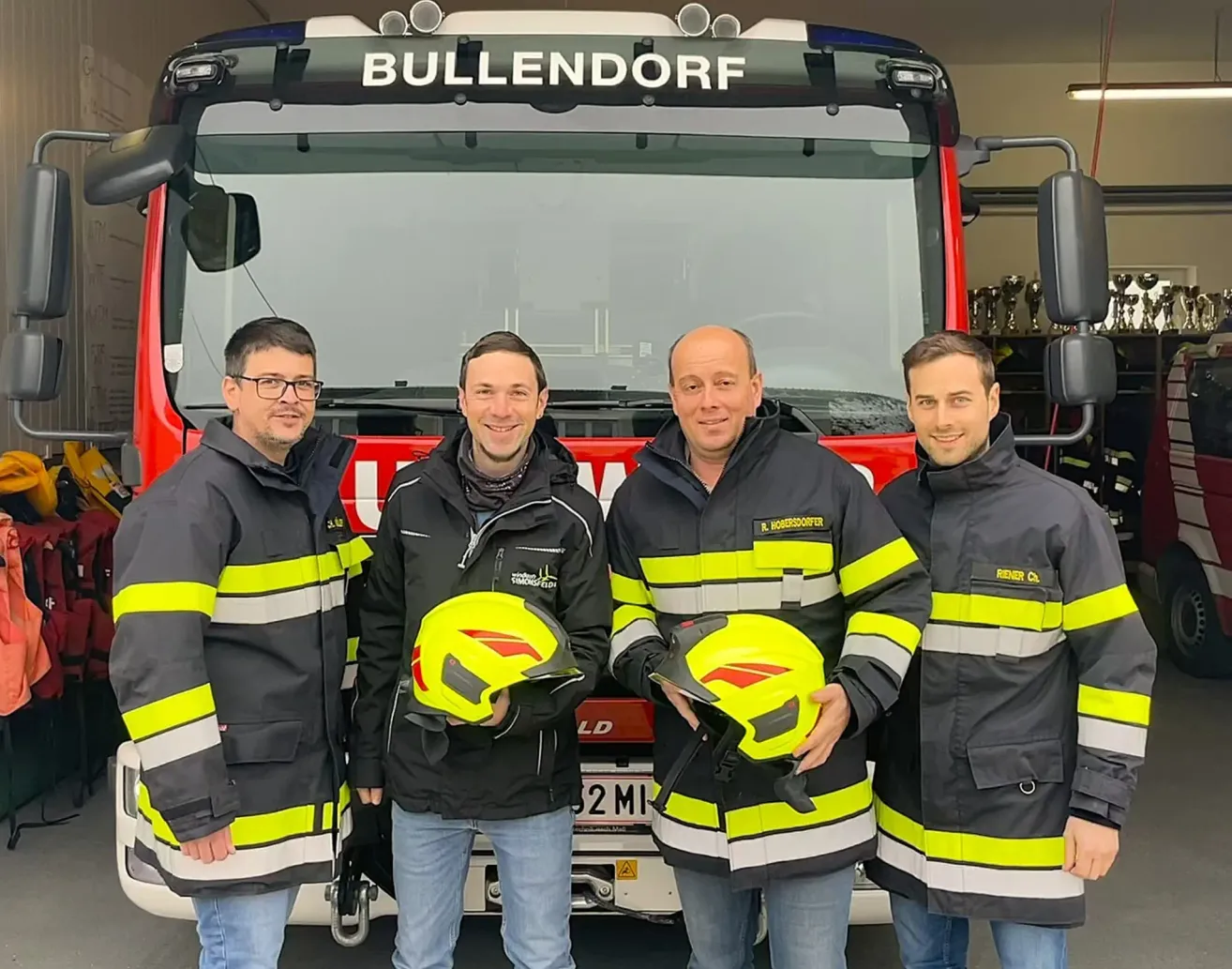 Vier Männer stehen vor einem Feuerwehrwagen. Drei von ihnen halten gelbe Helme. Der Wagen ist mit 'BULLENDORF' beschriftet. Die Männer tragen schwarze und gelbe Uniformen mit reflektierenden Streifen.