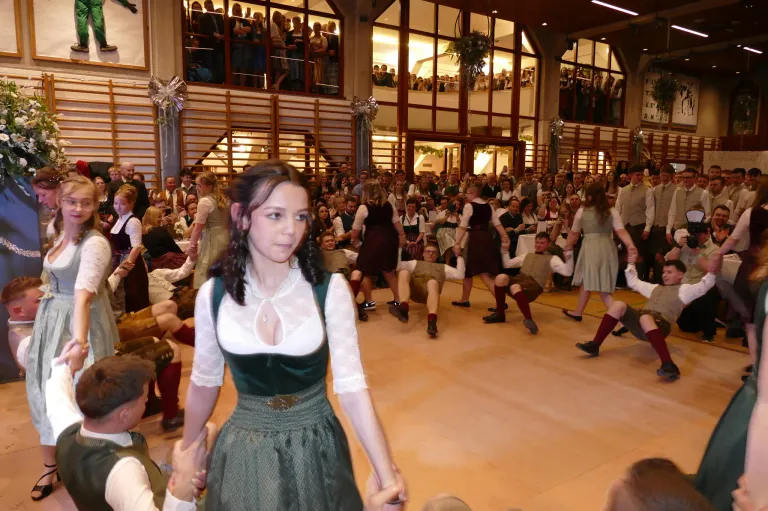 Ein großer Saal mit Zuschauern und Darstellern. Eine junge Frau in einem grünen Kleid tanzt mit einem Mann. Mehrere andere in traditioneller Kleidung tanzen in der Nähe.