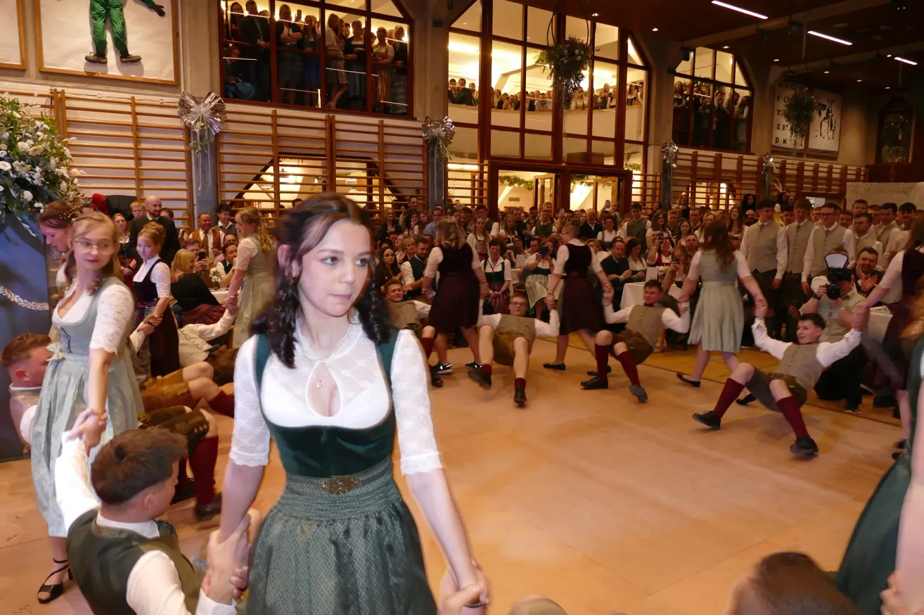 Ein großer Saal mit Zuschauern und Darstellern. Eine junge Frau in einem grünen Kleid tanzt mit einem Mann. Mehrere andere in traditioneller Kleidung tanzen in der Nähe.