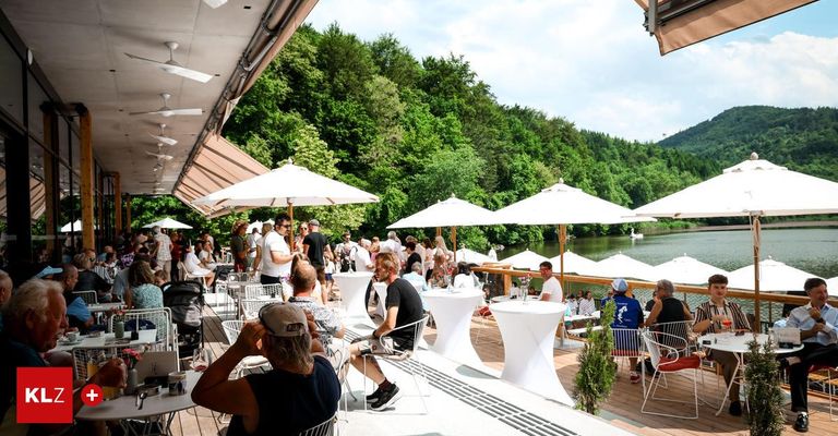 Eine Außenterrasse eines Cafés mit Blick auf einen See. Die Gäste sitzen unter Schirmen und genießen die Aussicht auf den See und die umliegende Grünfläche.
