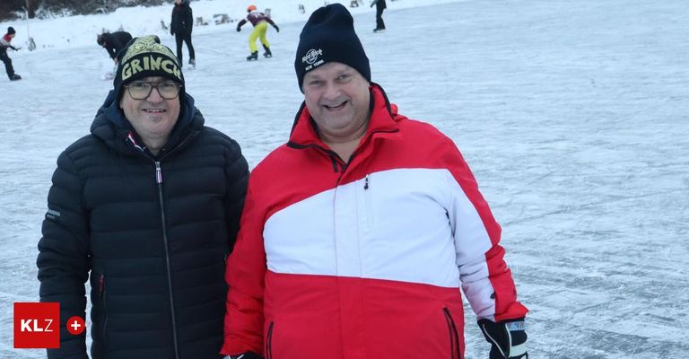Two men standing in the snow with a rink in the background. One man is wearing a red and white jacket.