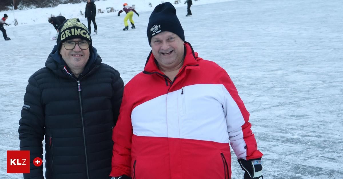 Two men standing in the snow with a rink in the background. One man is wearing a red and white jacket.