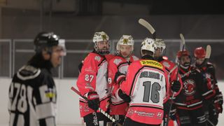 Eishockeyspieler versammeln sich auf der Eisbahn, tragen rote und weiße Trikots mit den Nummern 16, 87 und 9. Die Helme sind mit Logos verziert. Ein Spieler hält einen Eishockeyschläger.