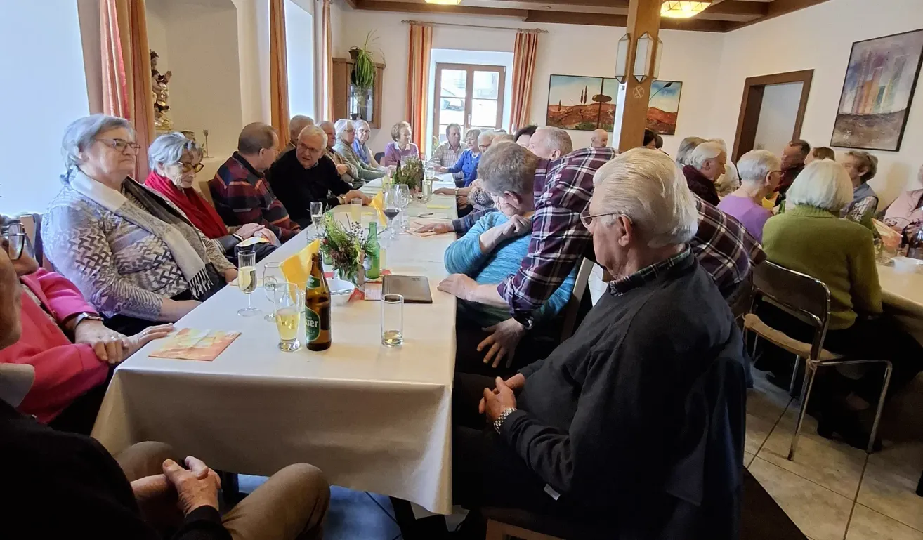 Eine Gruppe älterer Menschen versammelt sich um einen Tisch in einem Raum. Einige sitzen, andere stehen. Auf dem Tisch befinden sich Weingläser, eine Flasche und eine Blume.