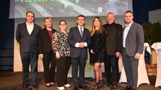 Eine Gruppe von Menschen steht auf einer Bühne und lächelt für ein Foto. Hinter ihnen zeigt eine große Leinwand Text. Der Text lautet 'freuen wir uns auf 2026.' Sie tragen formelle Kleidung, was auf eine formelle Veranstaltung hindeutet.