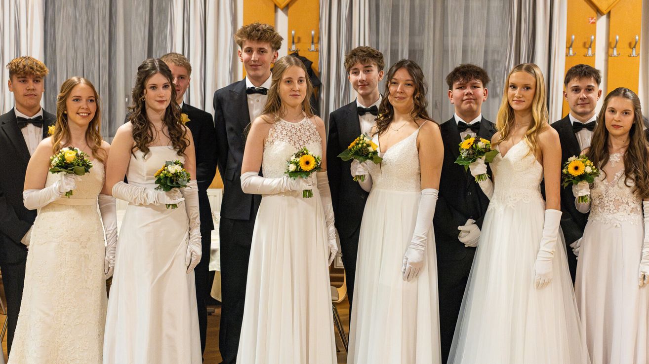 Eine Gruppe von Menschen, möglicherweise eine Hochzeitsfeier, steht in einem Raum mit weißen Vorhängen. Drei Frauen in weißen Kleidern halten Blumensträuße, und vier Männer in Anzügen stehen hinter ihnen. Sie tragen alle weiße Handschuhe.