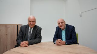 Zwei ältere Männer sitzen an einem Holztisch, lächelnd, einer mit einer gemusterten Krawatte, der andere in einem blauen Hemd und dunklem Anzug. Hinter ihnen eine schlichte weiße Wand.