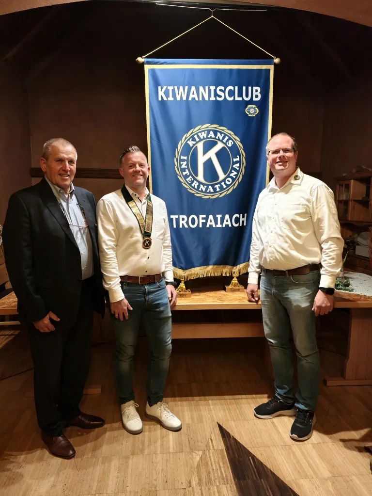 Three men stand in front of a blue banner with 'Kiwanisclub' and 'Kiwanis International' on it. One man wears a medal. Behind them is a wooden table with trophies.