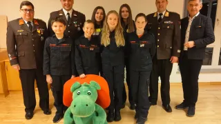 Eine Gruppe von Menschen in Uniformen mit Abzeichen steht um ein grünes Plüschtier auf einem Holzboden. Einige tragen Militäruniformen, andere Zivilkleidung. Eine Person trägt eine Brille.